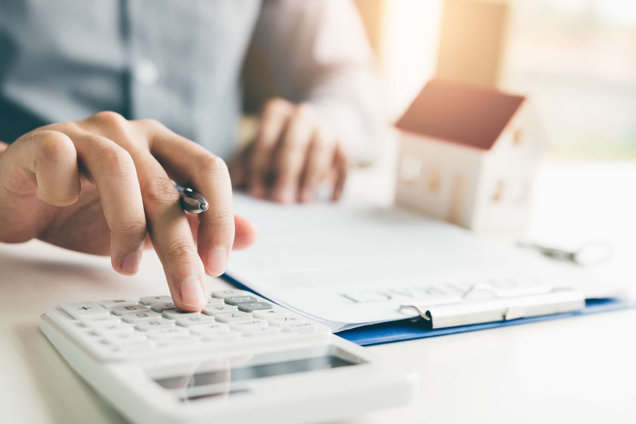 imagem com foco em uma mão de homem digitando em uma calculadora, com canetas, papeis e uma casa em miniatura do lado
